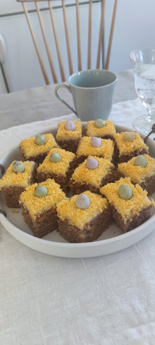 Fyrkantiga kakanbitar med gult strössel och påskägg, blå mugg och klart glas på linneduk.