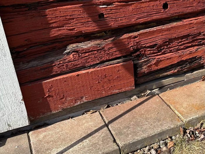 Nött röd trävägg, skugga, solbelyst trottoarkant, erosion, struktur, varma färger.