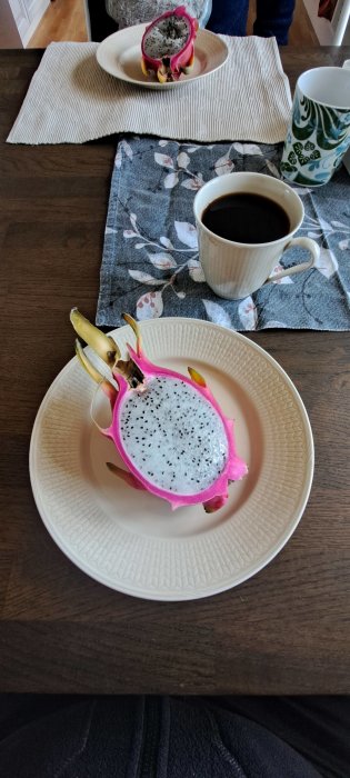 Frukostdukning med drakfrukt på tallrik och svart kaffe i kopp, över ett träbord.