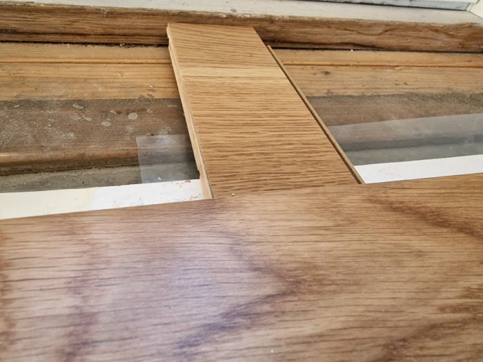 Två trägolvslameller ovanpå gammalt fönster, olik träfinish, ojämn övergång, renoveringsarbete pågår.