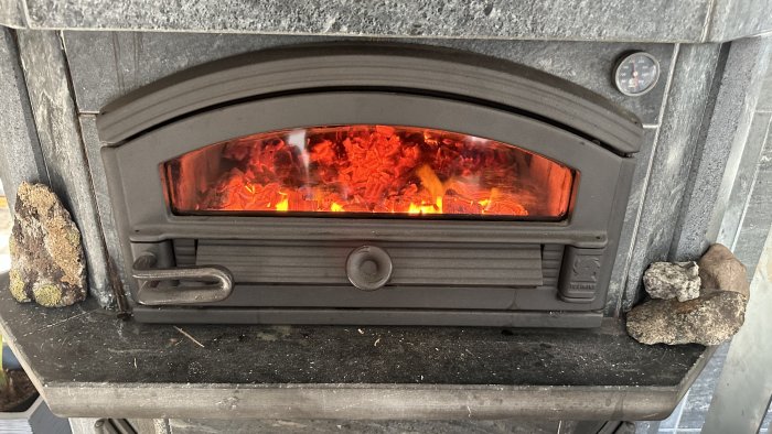 Brinnande ved i vedeldad kamin, stenar på sidorna, termometer visar hög temperatur.