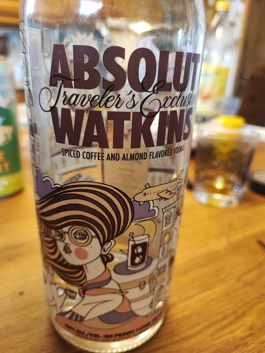 En vodkaflaska, "ABSOLUT Watkins", med kryddat kaffe och mandelsmak, illustrerad med en kvinnlig karaktär och resväska.