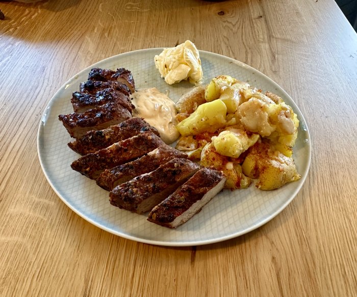 En tallrik med grillad kött, kryddade potatisar, sås och ett klump av smör.