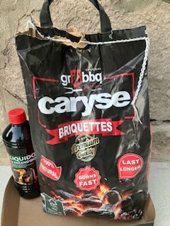 Svart kolpåse märkt "Caryse Briketter" bredvid en flaska grillvätska på kartong. Grilla, BBQ-tema.