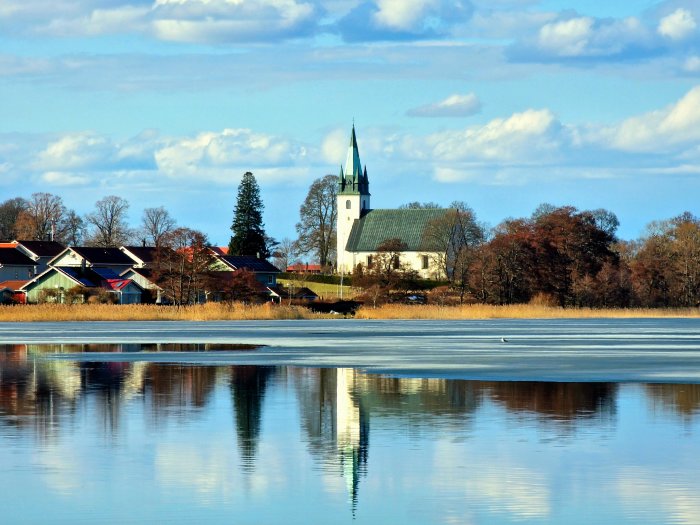 Landskapsvy, kyrka, hus, speglande vatten, isstrimmor, träd, klart väder, moln, lugnt, idylliskt, småstadskänsla.