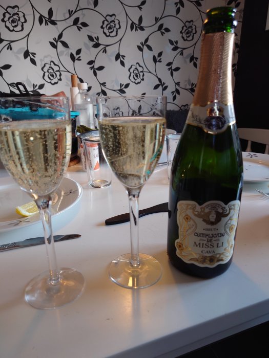 Två champagneglas, öppnad flaska cava, bakgrund med blommig tapet, desorienterat bordsläge, festlig eller social situation.