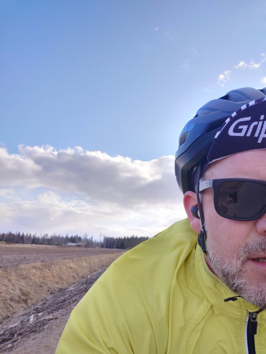 Cyklist med hjälm och solglasögon, gulfärgad jacka, landskap med öppet fält, molnig himmel.