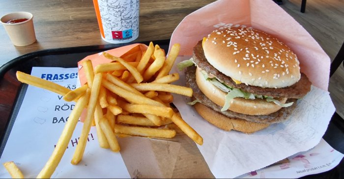 En dubbel cheeseburgare med pommes frites, ketchup och en läsk på ett serveringsbricka. Fastfood-rätt i en restaurangmiljö.