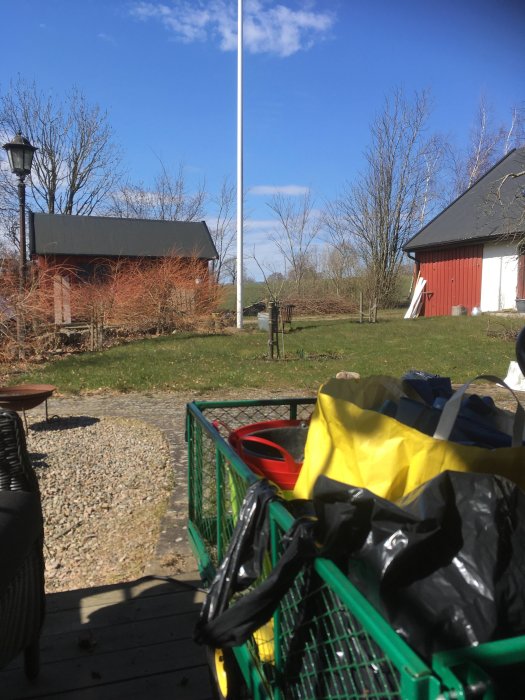 Utomhus med grönt kundvagn, gula och svarta påsar, trädgårdsmöbler, lantliga röda byggnader, flaggstång och blå himmel.