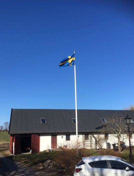 Svensk flagga vajar, klarblå himmel, lantlig byggnad, bilparkering.