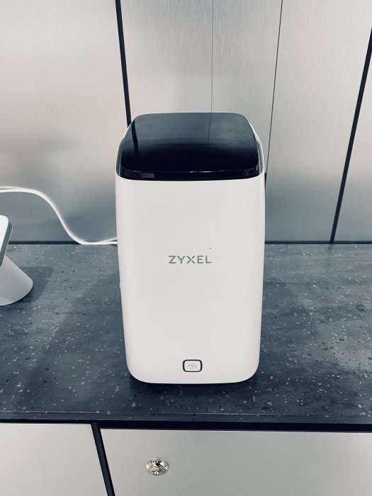 En vit Zyxel-router på en grå bänkskiva framför grå skåp med eluttag.