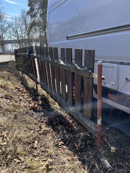Husvagn parkerad bakom trästaket längs med grusig mark nära natur på en solig dag.
