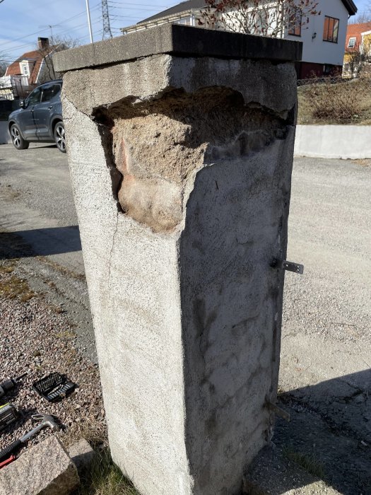 Skadad betongpelare med synlig armering, verktyg i förgrunden, bostadshus och bil i bakgrunden.