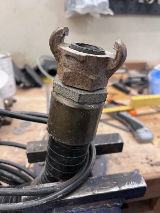 Bild på en rostig och sliten borrchuck mot en suddig verkstadsbakgrund, med verktyg och kablar.