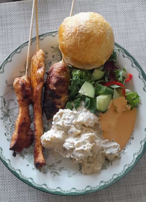 Grillad kyckling, bröd, sallad, potatissallad, och sås på en mönstrad tallrik.