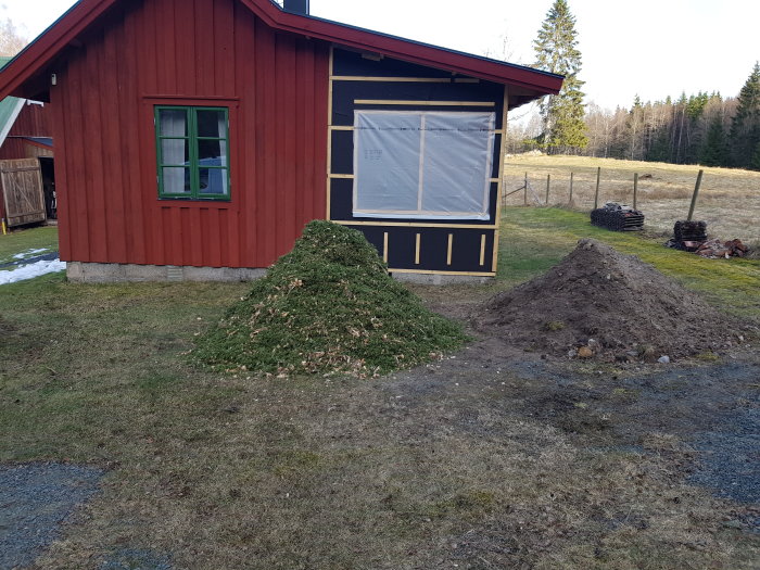 Röd stuga, grönt fönster, högar av grenar och jord, plasttäckt utbyggnad, smältande snö, landskap i bakgrunden.