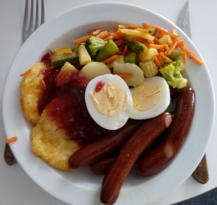 Tallrik med korv, potatis, ägg, grönsaker, brun sås på vit bakgrund.