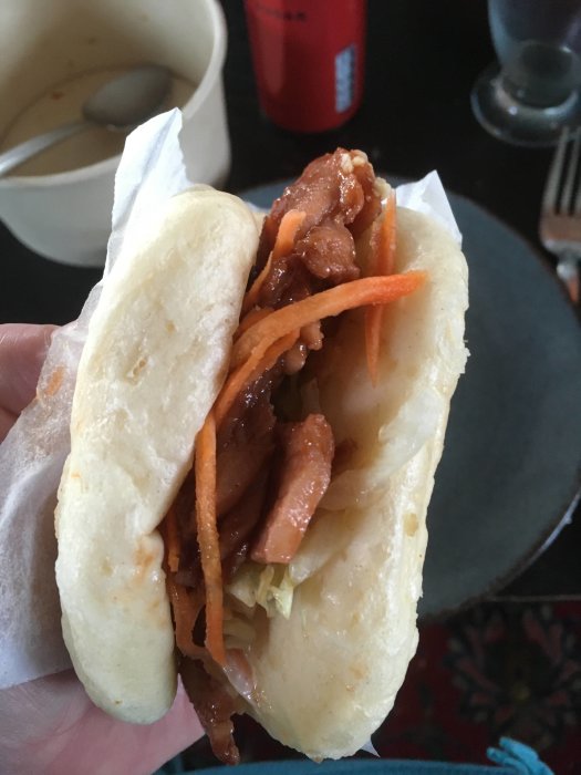 Närbild av en hand som håller en gua bao med fläsk och morötter, över en bord med en Coca-Cola burk.