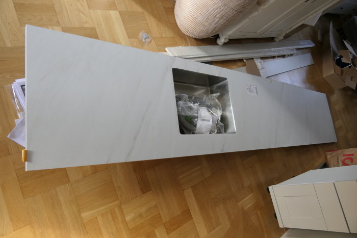 En omonterad möbeldel med en vit yta, ligger på ett parkettgolv, omgiven av kartonger och byggmaterial.