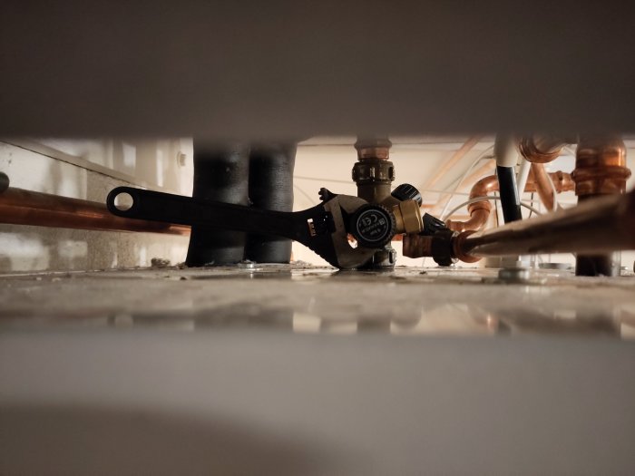 VVS-installation med kopparrör, kulventil och skiftnyckel på ett dammigt golv.