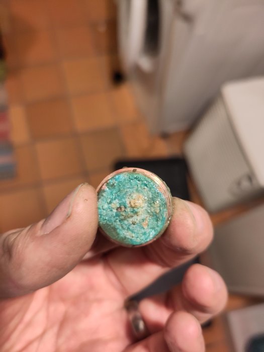 En hand håller ett föremål med blågrön beläggning eller korrosion, troligen en rör- eller vattenledningsdel.