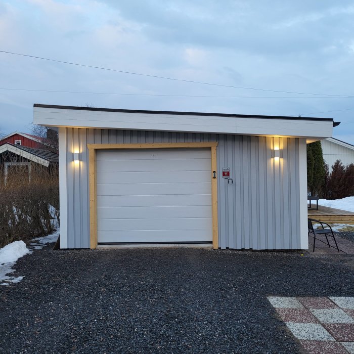En fristående garagebyggnad vid skymning med upplysta utomhuslampor och en vit garageport. Smältande snö syns.