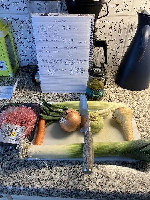 Förberedelse av mat med receptbok, grönsaker, köttfärs, och kryddburk på köksbänk.