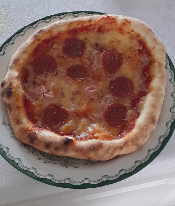 En pizza med salami och skinka på en mönstrad tallrik, ser nybakad och varm ut.