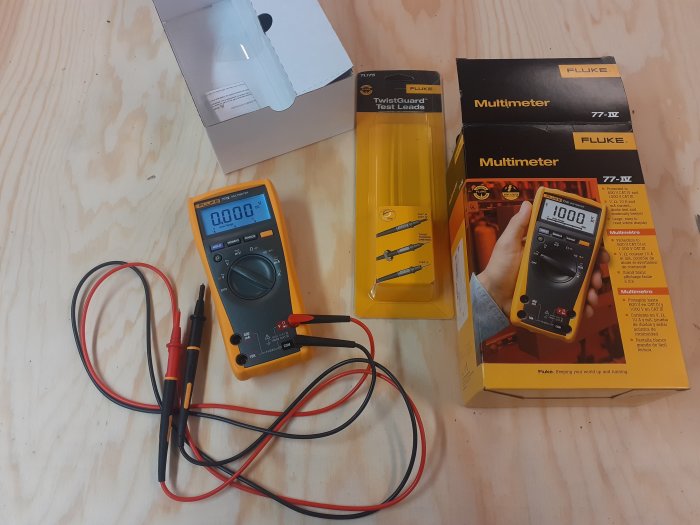 Digital multimeter med testledningar bredvid dess förpackning och en extra förpackning av testledningar på träbänk.