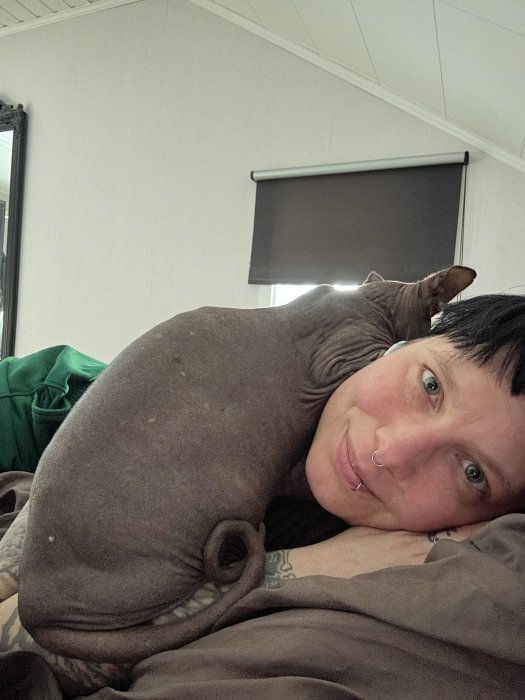En person med en näspiercing och en Sphynx-katt vilande på dem. Intima, mysiga, inne, säng, tatuering, hårdför, kärleksfull blick.