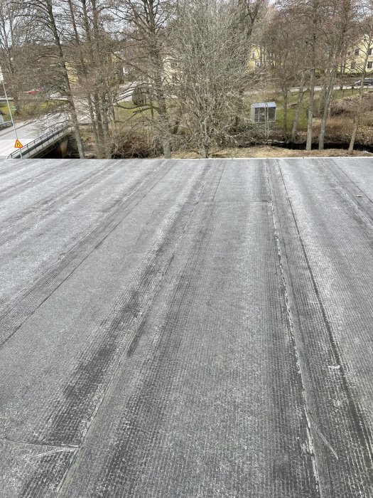 Tak med svarta takbeläggningsskivor, träbro, träd utan löv, byggnader i bakgrunden, dagtid, molnig himmel.