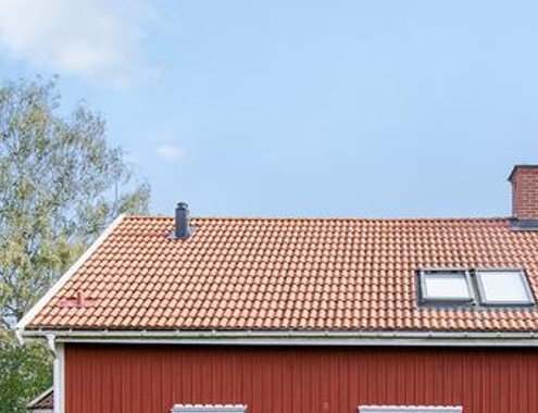 Ett rött trähus med tegeltak, två vindsvåningsfönster, delvis dolt av grönska under en blå himmel.