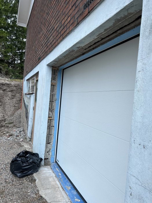 Garage med vit dörr, tegelfasad, pågående konstruktionsarbete, svart sopsäck, ofärdigt yttre.