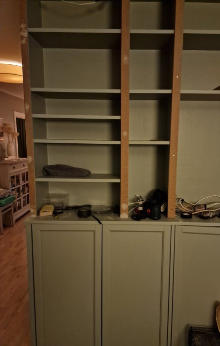 Ett ofärdigt bokhyllskåp med tomma hyllplan och diverse föremål på skranket i ett hemmiljö.