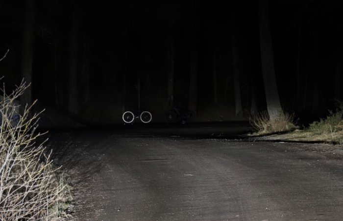 Mörk skogsväg vid natt, upplyst av fordon, med parkerad cykel i bakgrunden.