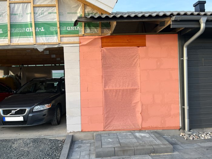 Renovering av hus, rosa isoleringsskiva, grå bil, delvis öppen carport, byggmaterial synliga, stenplattor förgrund.