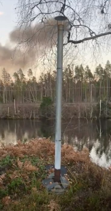 Metallrör vid en sjö med skog i bakgrunden och brun vegetation nära, möjlig rök eller smuts i luften.