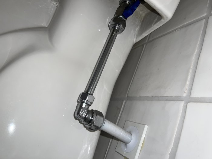 Toalettens insida med metallanslutning och vita kakelväggar. Regelverk och stadighet synes. Hygien- eller reparationssammanhang möjligt.