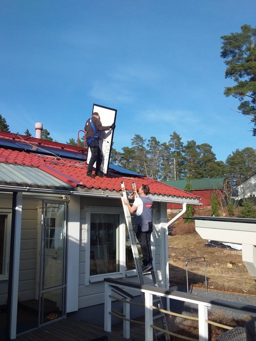 Två personer monterar fönster på hus. Blå himmel, röd hustak, träveranda. Arbete pågår, säkerhetsåtgärder syns.