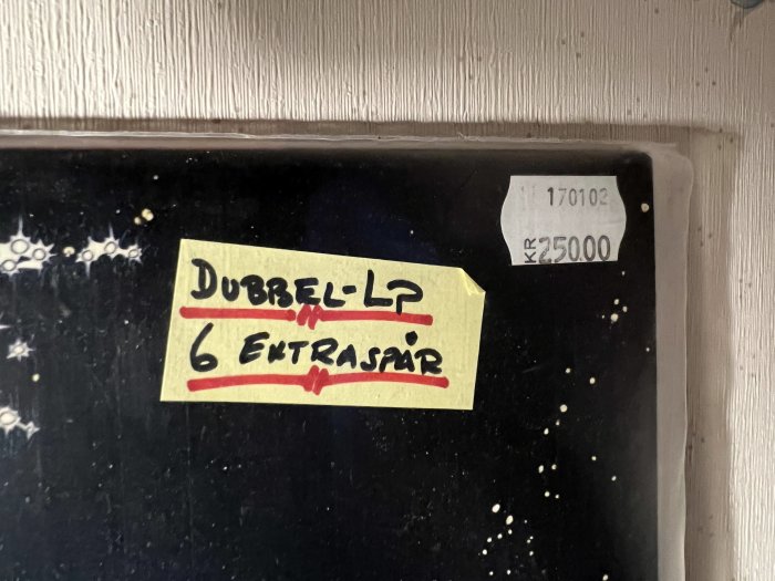 Lapp med text "DUBBEL-LP 6 EXTRASPÅR", prisinformation, mot suddig svart bakgrund.