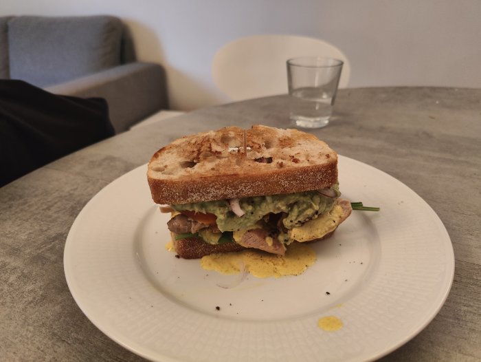 En smörgås på en vit tallrik, mörkt bröd, grönsaker, sovsdroppar, bakgrund med soffa och vattenglas.