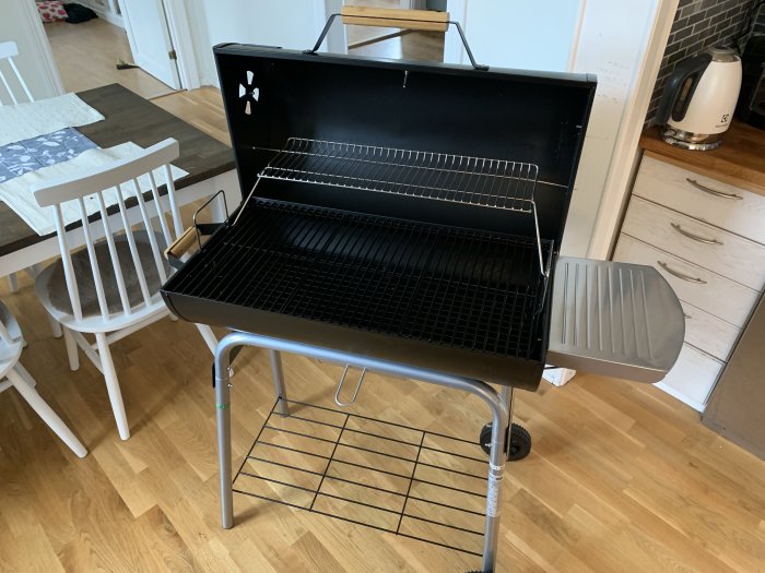 En svart kolgrill med hjul inne i ett kök, ej i bruk.