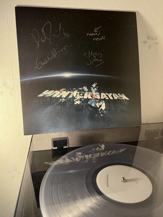 Vinylskiva på spelare, albumomslag med autografer, rymdtema, bordskonstellation med människor och instrument på jorden.