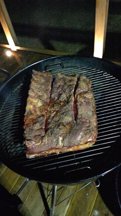 Stor revbensspjäll på kolgrill om natten, belysning i bakgrunden, matlagningsaktivitet.