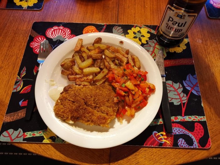 Måltid med schnitzel, rostad potatis, salsa, majonnäs och öl på en mönstrad bordstablett.