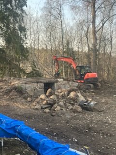 En orange grävmaskin vid skogsområde, arbetar bland nedfallna träd och stenar, med blå plast presenning i förgrunden.