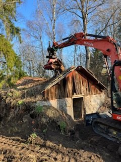 Grävmaskin vid markarbete nära en liten byggnad med naturbakgrund under klar himmel.