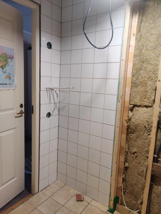 Renovering pågår, öppen vägg med isolering, vitt kakel, dusch slang, världskarta, vit dörr.