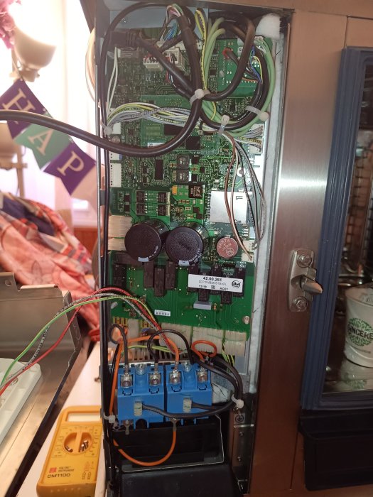 Ett öppet elektroniskt kabinett med kretskort, kablar, kondensatorer och en multimeter på sidan.