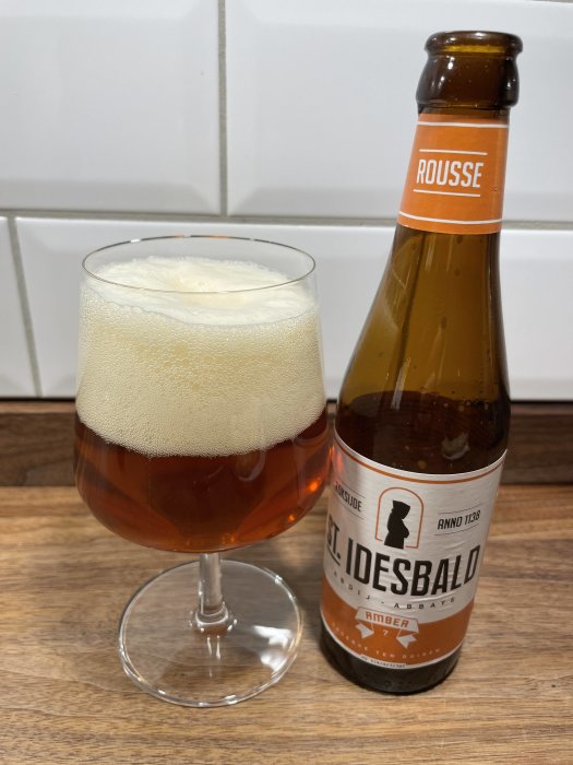Ölglas med skum, flaska märkt "Rousse" och "St. Idesbald", bänk och kakelvägg.